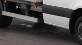 Side view of a white truck cab and trailer parked on a paved surface; a long side step is visible beneath the cab and a circular manhole cover with an X lies on the ground beneath the truck.