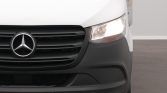 Close-up of a white Mercedes van's front end, showing the grille, emblem, and a lit headlight on the right side.