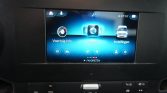 Car infotainment touchscreen showing Voertuig Info, Apps, Instellingen options with a blue UI backdrop and row of icons at the bottom
