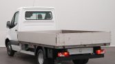 White Mercedes Sprinter flatbed truck seen from the rear-left side, empty cargo bed and rear lights visible in a neutral setting