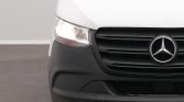 Front view of a white Mercedes van, showing the black grille and circular Mercedes emblem center stage with a headlight on the left side.