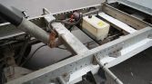 Close-up of a truck trailer chassis showing a hydraulic arm, wiring, and a small white battery box with a yellow warning label,