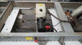 Electric pump attached to a metal trailer frame with a translucent reservoir, hoses, and wiring; appears to be a small fluid pump assembly on equipment.