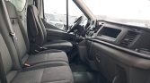 Front cabin of a dark van with driver and passenger seats, steering wheel, gear shifter, and a wide dashboard viewed from the passenger side exterior vehicles visible outside the windows.