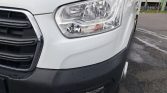 Front end of a white van showing the large headlight, chrome trim, and black bumper on a wet pavement surface