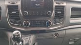 Interior view of a vehicle dashboard: center infotainment screen, radio controls, and climate dials with a six-speed manual gear stick in the foreground.