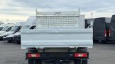 Rear view of a light gray flatbed truck with a perforated metal headboard, parked among white vans in a lot.