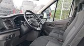 Interior of a white cargo van: driver and front passenger seats, steering wheel, and dashboard visible from the passenger side window. Outside, other vans are seen through the windshield on a cloudy day.