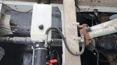 White plastic hydraulic fluid reservoir connected to a black pump, mounted on a rusted truck frame with hoses and wires nearby.