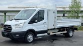 White Ford transit flatbed truck parked outdoors, with a tall metal storage box behind the cab and a long open cargo bed.
