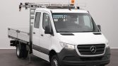 White Mercedes-Benz utility truck with a metal ladder rack on top and an orange beacon, indoors in a studio setting.