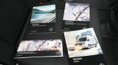 Four Mercedes‑Benz manuals (Sprinter and maintenance guides) laid on a dark car seat. Graphics show road, wheel logo, and a van image.