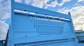 Blue metal truck tailgate with a perforated vent panel and tied ropes, set against a blue sky with clouds.