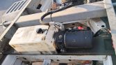 Close-up of a dusty hydraulic pump and motor mounted on a metal frame with hoses and a white reservoir tank nearby.