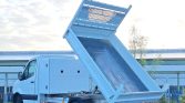 White utility van with a raised blue dump bed for unloading material in a parking lot or industrial area