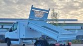 White Mercedes-style utility truck with a blue hydraulic dump bed raised for unloading in a parking lot.