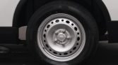 Close-up of a car wheel: a silver steel rim with circular holes and lug nuts on a black tire toward a white vehicle body edge