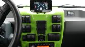 Bright green vehicle dashboard with a central tablet-like display mounted on top, steering wheel to the left, and numerous black switches and controls below.