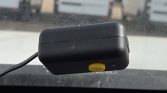 Windshield-mounted black rectangular device with a yellow circular button, connected by a cable, seen from inside a car.