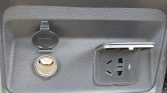 Open 12V cigarette-lighter socket on a car panel beside a hinged-cover AC power outlet.