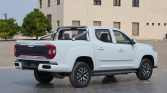 White double-cab Maxus pickup with a metal roll bar in the bed, parked on a tiled surface near a beige building.