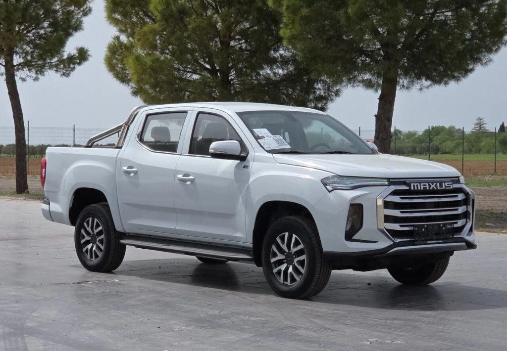 White Maxus pickup truck parked outdoors on a paved lot with trees in the background, front three-quarter view.