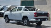 White pickup truck with a metal roll bar parked in a sales lot beside white vans against a beige building.