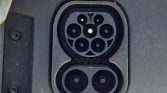 Close-up of an electric vehicle charging inlet with multiple round contacts: two large lower sockets and a circular cluster of five smaller sockets in a recessed panel on a dark car body.