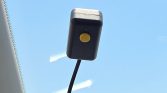 Black rectangular device mounted to the windshield with a yellow circular button in the center and a cord trailing downward into the car interior.