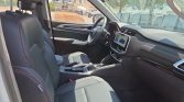 Front passenger and driver seating in a modern car with black leather seats and red contrast stitching, center console, and a touchscreen infotainment display on the dash showing a blue car image at a sunny day showroom lot outside the window.