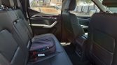 Back seat of a dark leather car interior with a gray backpack on the left rear seat; red stitching, sunny day outside shows trucks.