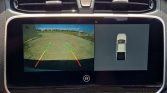 Car infotainment screen showing rearview camera with guiding lines (green, yellow, red) and a top-down vehicle view for parking help, text in Spanish warning to check surroundings.