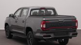 Rear three-quarter view of a gray Maxus pickup truck with a black tonneau cover and bold MAXUS tailgate lettering in a showroom setting