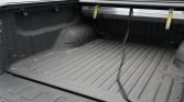Empty pickup truck bed with a ribbed, charcoal bed liner; two yellow strap anchors along the upper edge and a black strap draped across the bed.