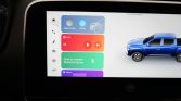 Infotainment screen showing colorful app tiles (Apple CarPlay-style) with a blue pickup truck graphic on the right side of the display in a car interior scene.