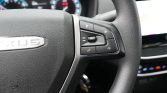 Close-up of a car steering wheel showing control buttons on the right (volume, source, phone, mute) with a blurred dashboard in the background.