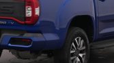 Blue pickup truck viewed from the rear driver's side, showing the taillight, bumper, and wheel on a dark pavement surface.