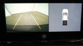 Rearview camera feed with guiding lines and orange markers, showing a clear parking area behind the vehicle, left side of the display.