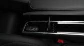 Center console in a dark car interior with two USB ports (USB-A and USB-C) and a wireless charging pad, accented by chrome trim and black leather with white stitching. A red seatbelt buckle is visible nearby.