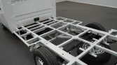 White truck cab with the bed removed, exposing a silver metal frame chassis and attached wheels, indoors on a gray floor.