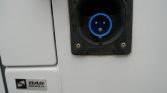 Blue circular 5-pin power inlet mounted on a white vehicle near a BAS World badge, industrial trailer connector close-up.