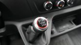 Close-up of a manual transmission gear shift knob showing R and gears 1–6 with a red accent, dashboard controls blurred in the background.