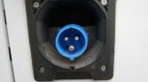 Blue IEC 60309 power inlet (3-pin) recessed in a black panel on a white surface, used for outdoor/industrial power connection