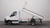 White flatbed/dump truck shown in profile with its bed raised, ready to unload materials in an empty lot.