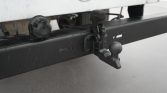 Close-up of a trailer hitch on a black bumper: a tow ball and mounting bracket, with a circular electrical connector nearby under a white vehicle body.
