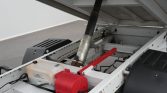 Open equipment bay with a hydraulic actuator, red latch, and a white reservoir inside a metal frame.