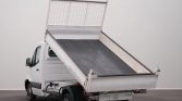 White light-duty dump truck with its cargo bed raised, ready to unload on a plain indoor backdrop.