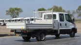 Rear three-quarter view of a white flatbed utility truck with a metal mesh headboard, parked in a lot with other trucks nearby.