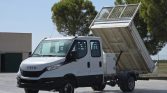 White Iveco dump truck with its bed raised for dumping, parked on a paved lot with trees in the background