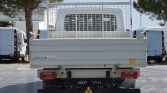 Rear view of a white flatbed truck with a metal mesh cargo barrier, parked in a lot with white vans nearby and trees in the background.